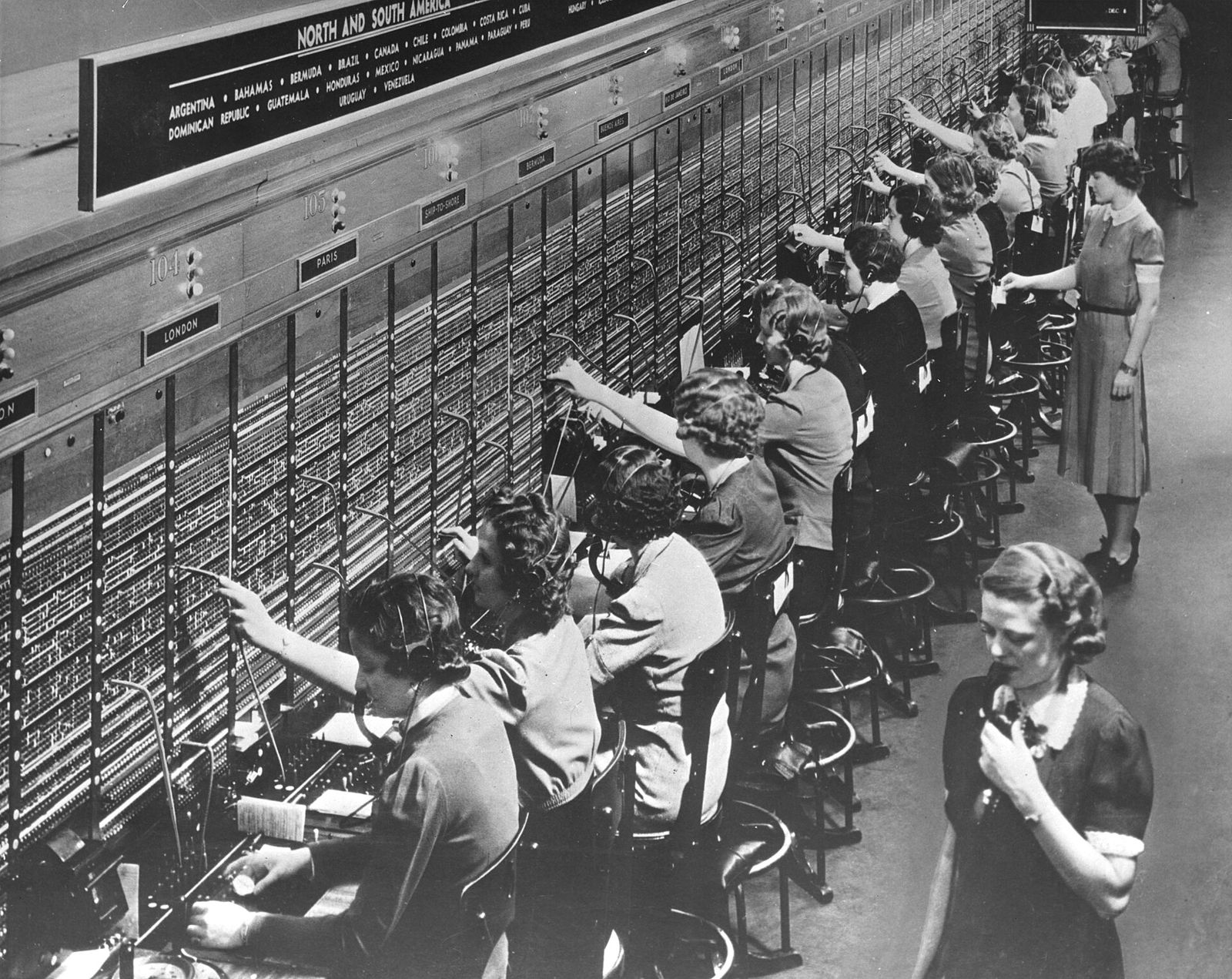 Una fila de telefonistas conecta llamadas a mano en una gran centralita, con auriculares y cables.